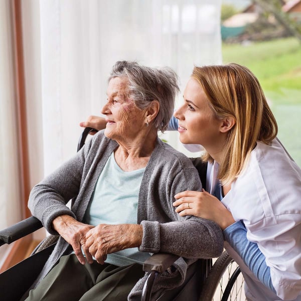 A nurse Neals near a senior as she look's out a window.